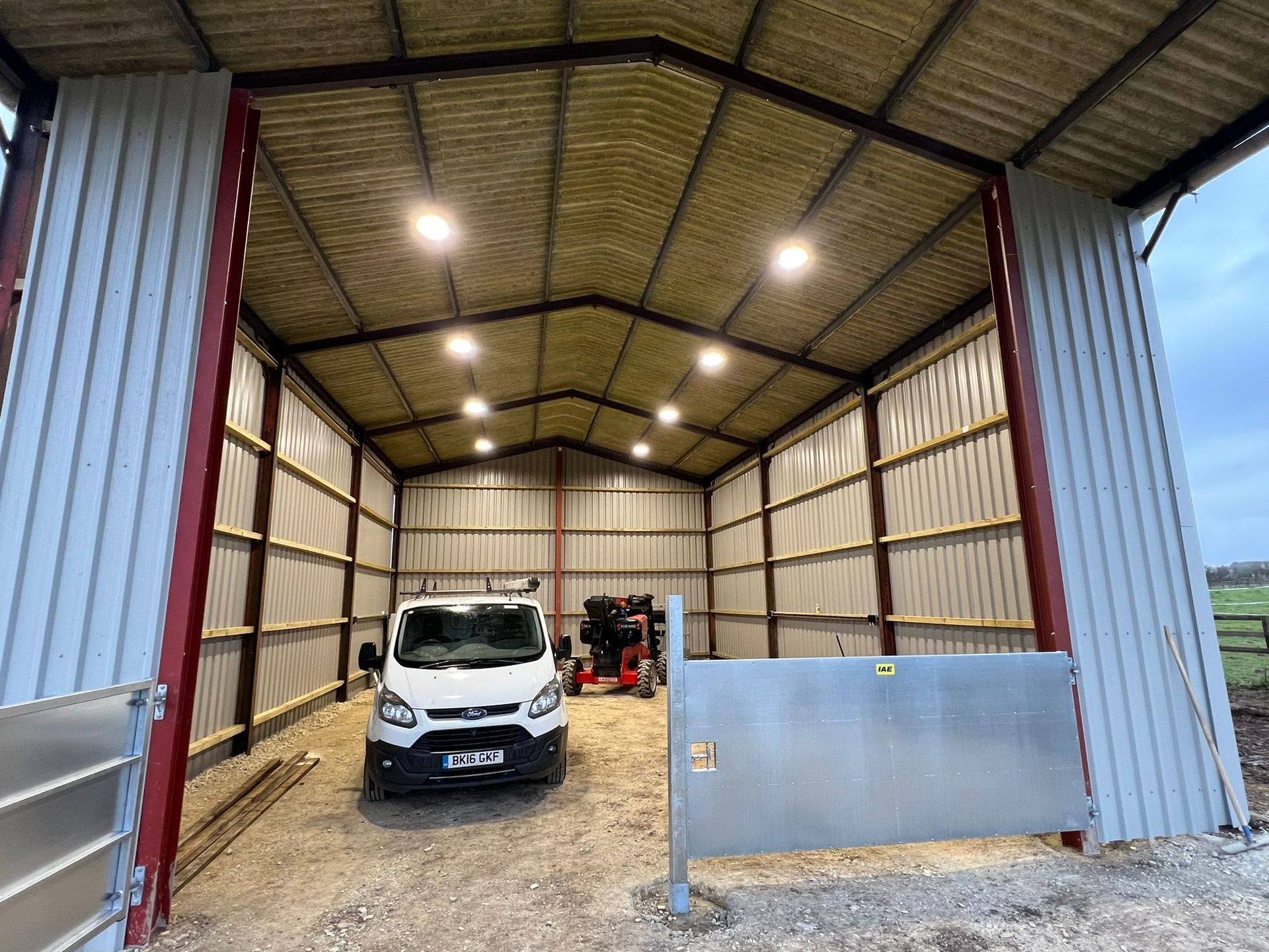 A white van parked inside a large, open-ended metal agricultural shed with overhead lighting.