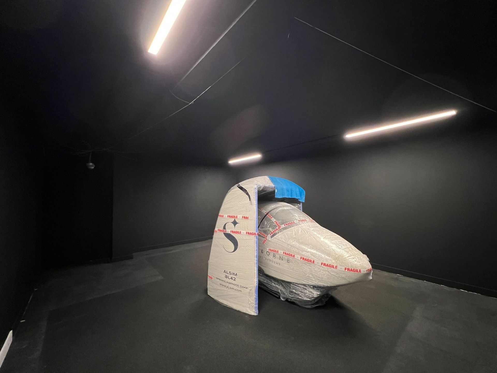An airplane cockpit section sits wrapped in plastic and tape inside a minimalist, black-walled gallery space.