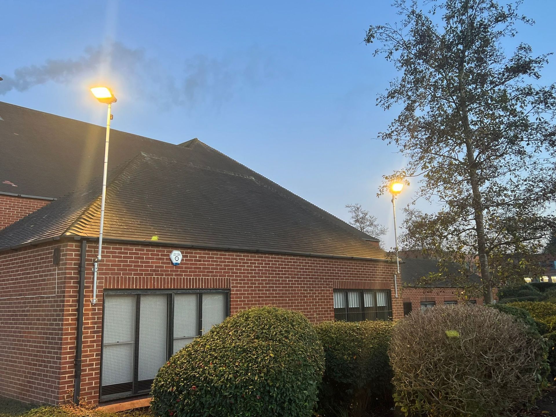 A brick building at dusk with two illuminated outdoor lamp posts and rounded shrubs in front.