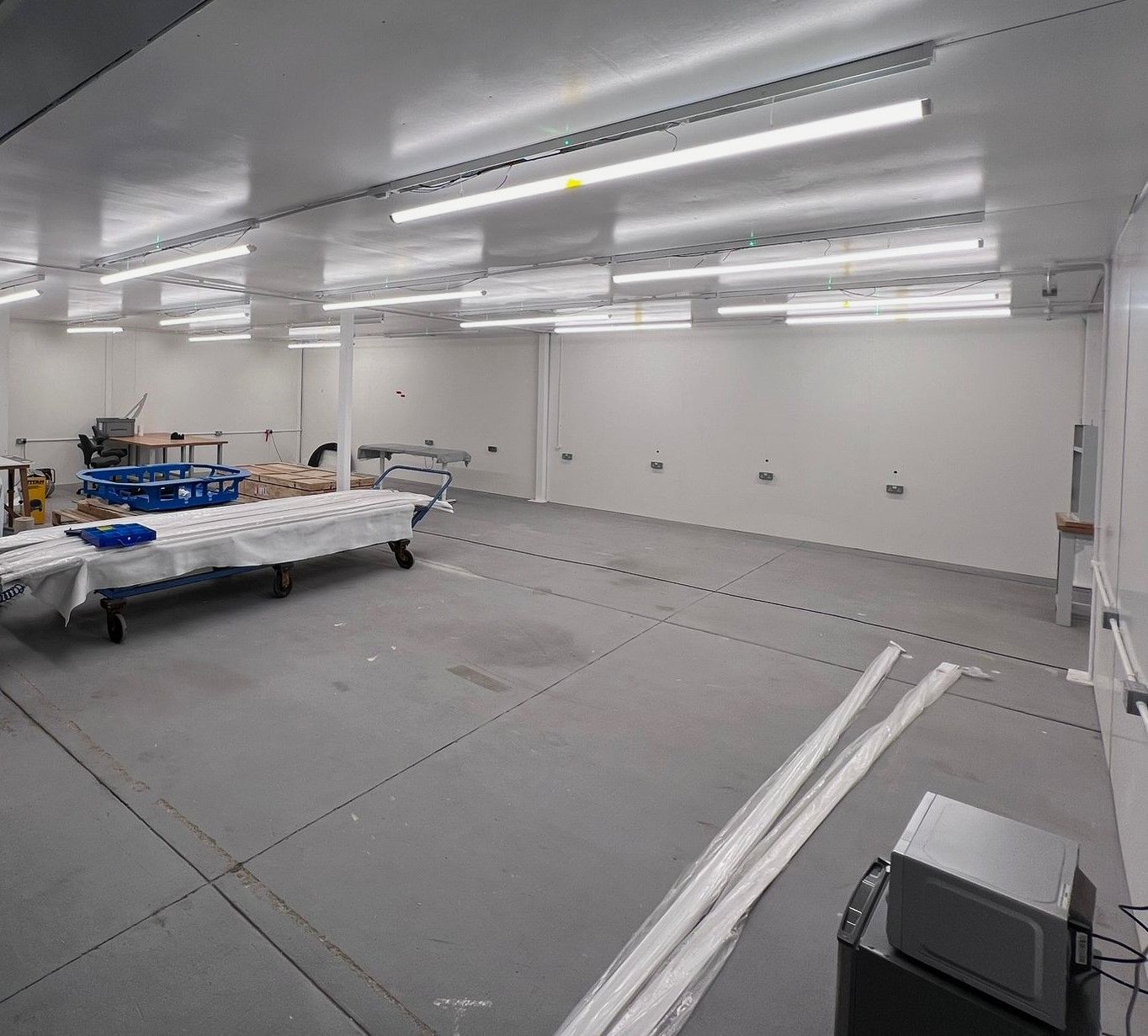 A large, empty white industrial room with bright overhead fluorescent lights, a rolling table, and a grey floor.