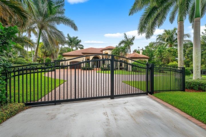 A black iron driveway gate in front of a sprawling, single-story house surrounded by palm trees and lush green lawns.