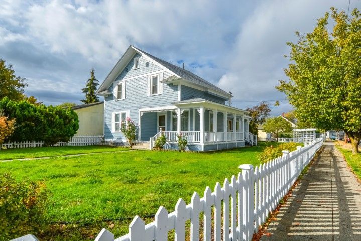 A light blue two-story farmhouse with a white front porch behind a picket fence along a sidewalk on a sunny day.