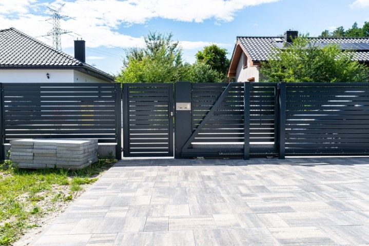 A modern black horizontal slat fence with a matching gate and pedestrian door, set on a paved driveway near suburban homes.