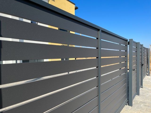 A modern, charcoal-gray horizontal slat privacy fence stands next to a paved walkway on a sunny day.