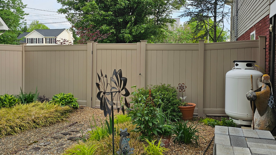A tan vinyl privacy fence borders a backyard garden featuring various plants, a metal garden spinner, and a propane tank.