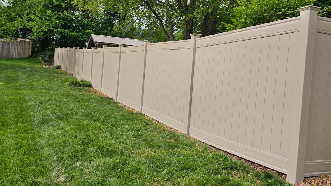 A tan vinyl privacy fence stretches across a green grass lawn under trees.