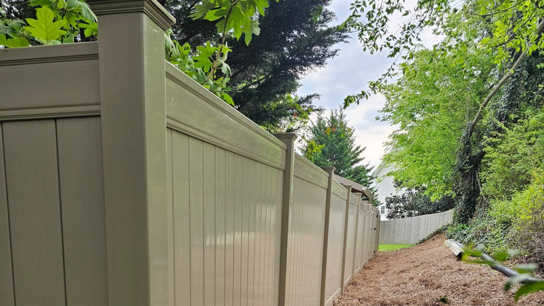 A tan vinyl privacy fence runs alongside a dirt path bordered by lush green trees and foliage.