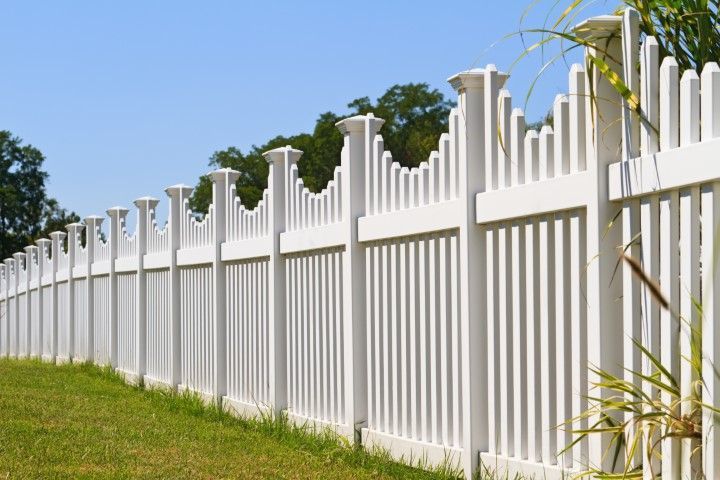 A long, white vinyl scalloped-top picket fence lines a green grassy lawn under a clear blue sky.