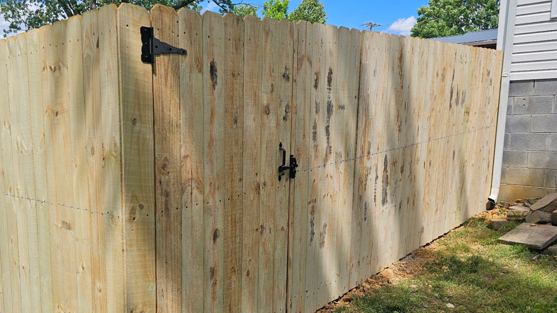 A wooden privacy fence gate with black metal hinges and a latch, set against a house exterior on a sunny day.