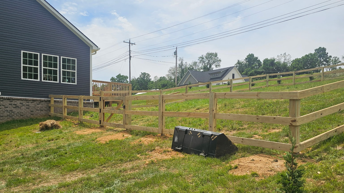 A dark blue house stands next to a wooden fence on a sloping, grassy lot on a sunny day.