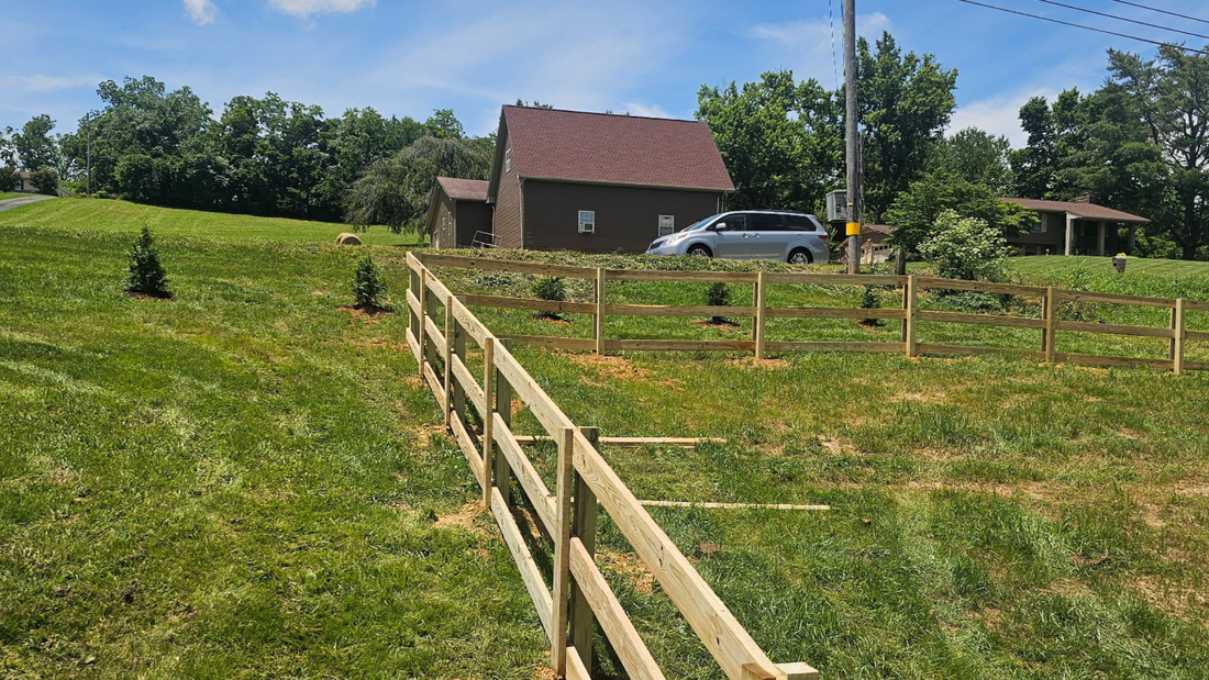 A wooden split-rail fence runs across a grassy field leading toward a brown house and silver van on a sunny day.