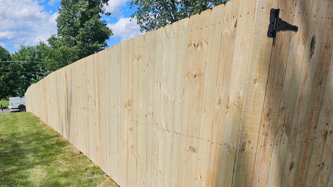 A long wooden privacy fence made of plywood sheets stands in a grassy area under a sunny, blue, partly cloudy sky.