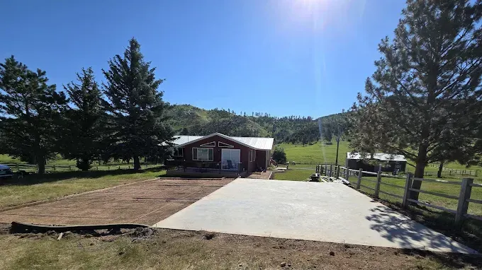 Red-sided cabin with concrete driveway, surrounded by trees and hills on a sunny day.