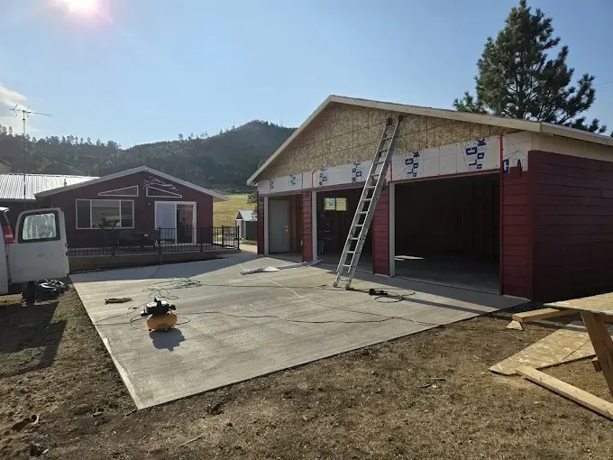 Garage under construction next to a red house, both in a rural setting. Ladder leans against the garage.