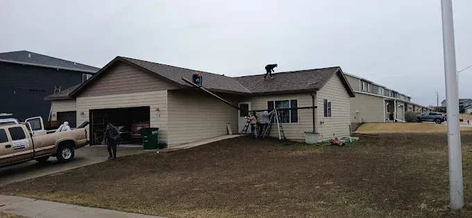House being worked on with people and truck. Garage door open, brown roof, cloudy day.