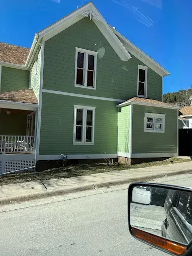 Green two-story house with white trim and windows on a sunny street.
