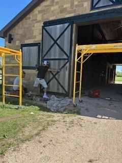 Worker painting a large barn door black and white beside yellow scaffolding and an open garage.