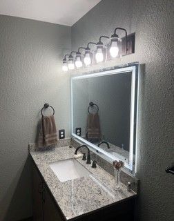 Modern bathroom vanity with granite counter, large lit mirror, sink, and wall sconce lights.