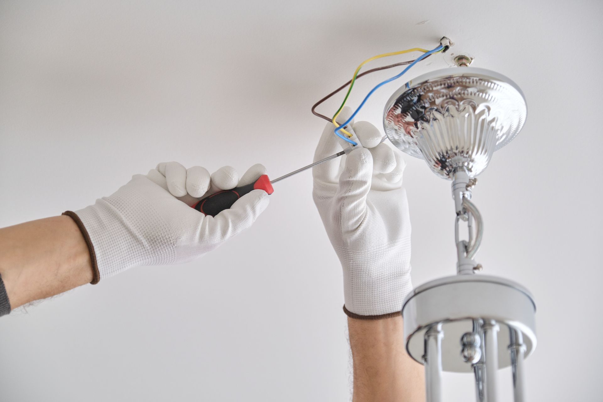 Hands in white gloves adjusting wires on a ceiling light fixture with a screwdriver.