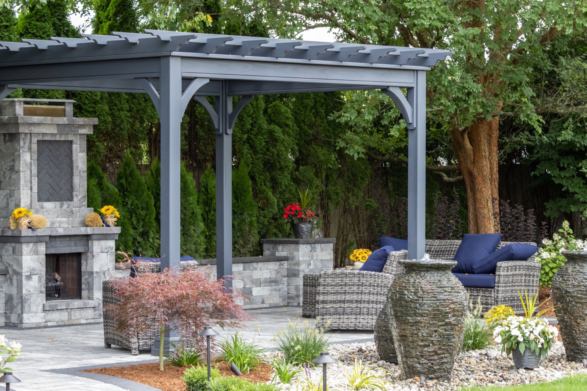 Outdoor patio with gray pergola, stone fireplace, and blue seating surrounded by lush greenery