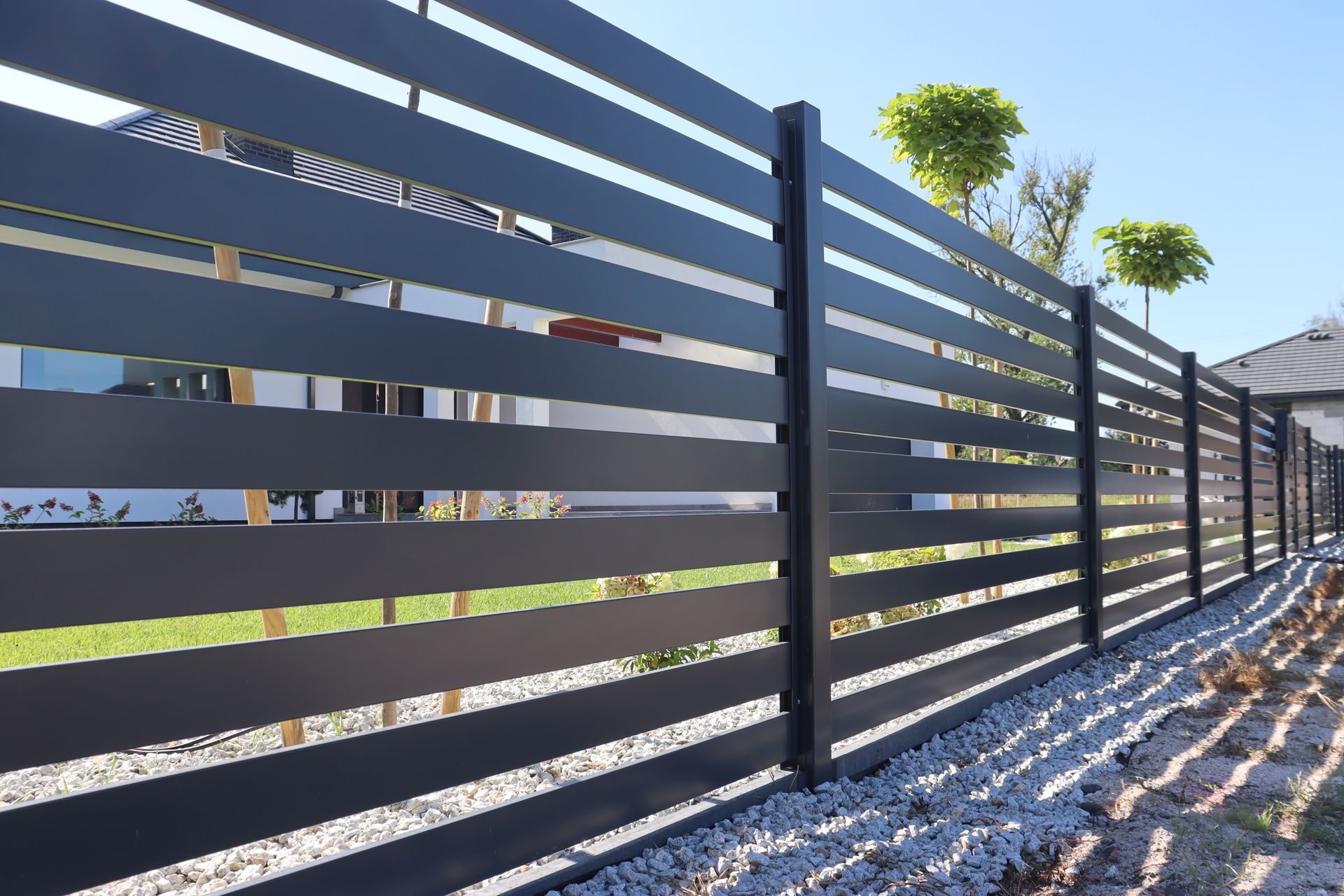 Modern gray slatted metal fence beside a gravel path, with sunlight and sparse trees in the background