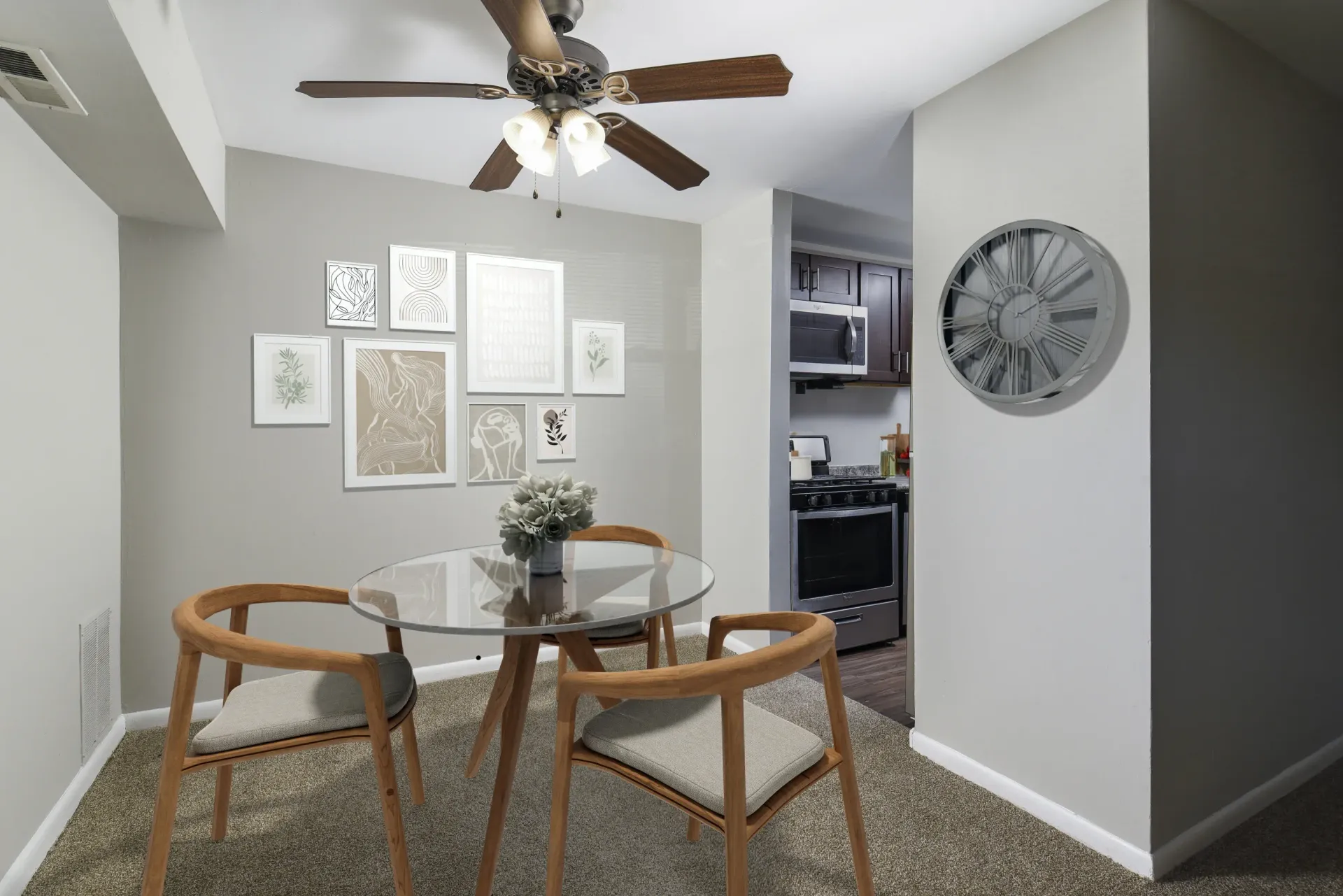 Dining area with a glass table, three chairs, and a gallery wall. A fan hangs from the ceiling. Kitchen in background.