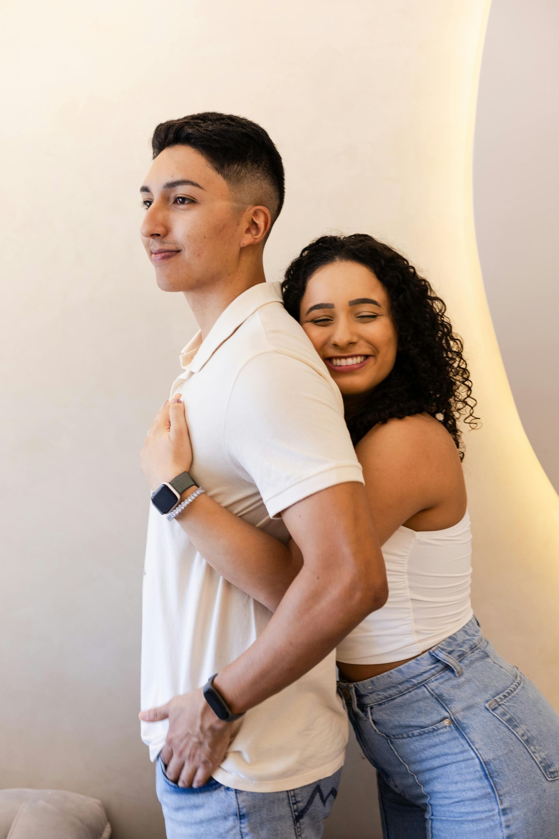 Woman hugging a person from behind. Both are smiling. Light-colored, neutral background.
