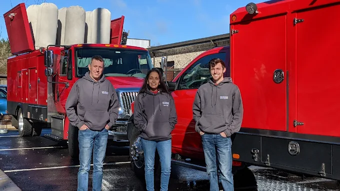 Three people stand in front of red trucks. Two are wearing grey hoodies and jeans. The third is wearing a grey hoodie and jeans.