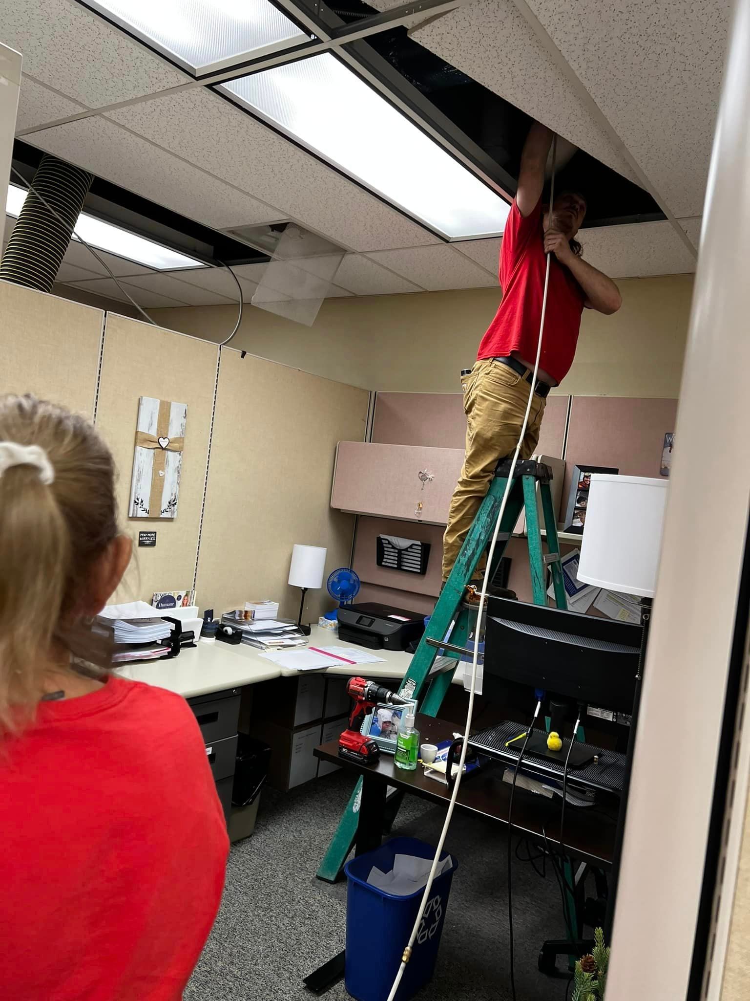 Person on ladder, working on ceiling tiles in office. A second person looks on.