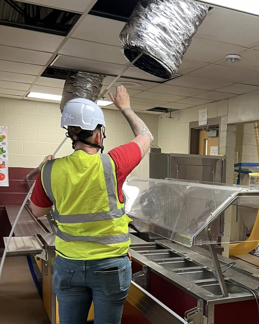 A worker in a hard hat and high-visibility vest pulls a flexible vent hose from a ceiling grid above a food service line.