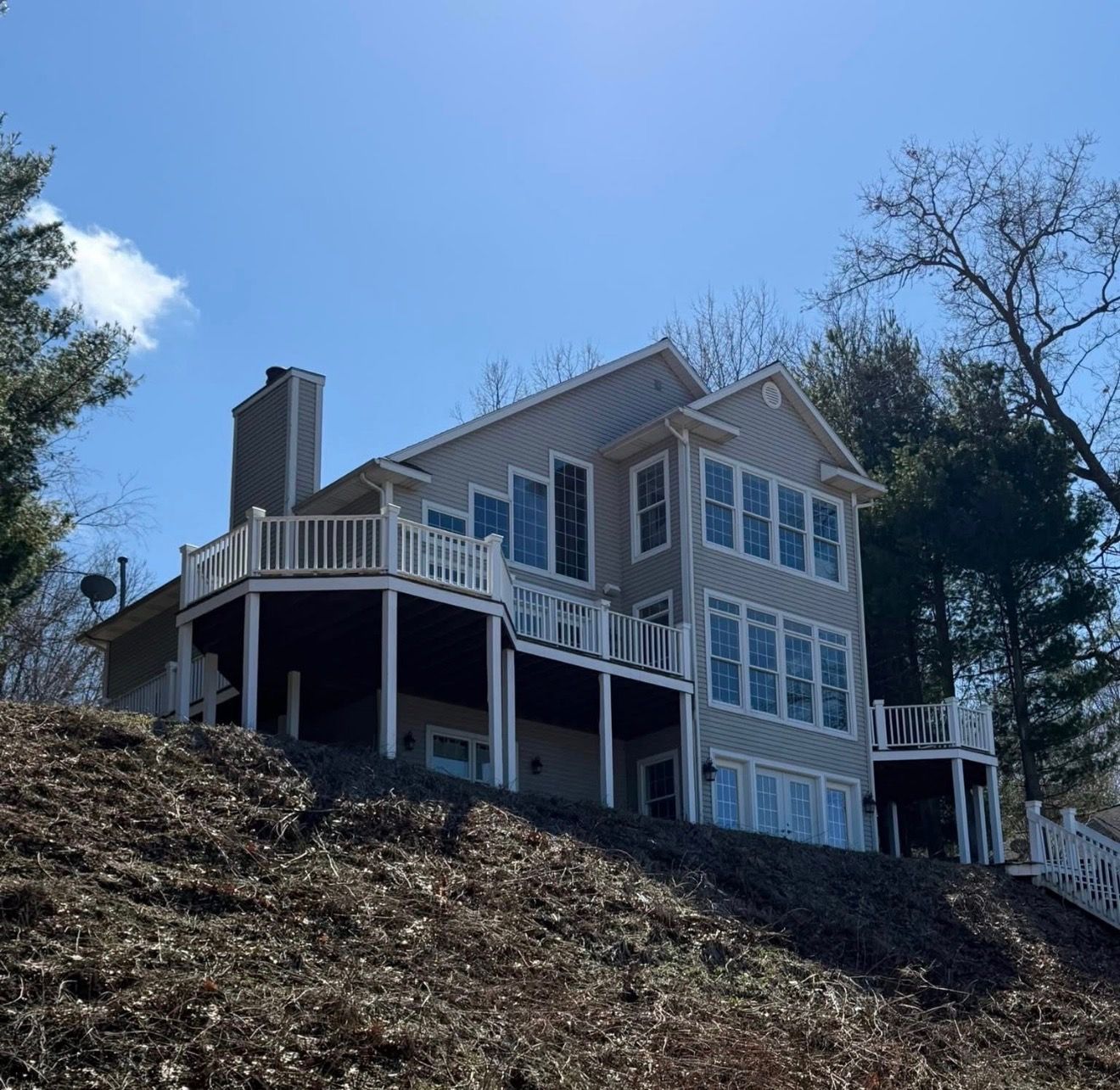 Large gray house with white trim and wraparound deck on a wooded hillside under a blue sky