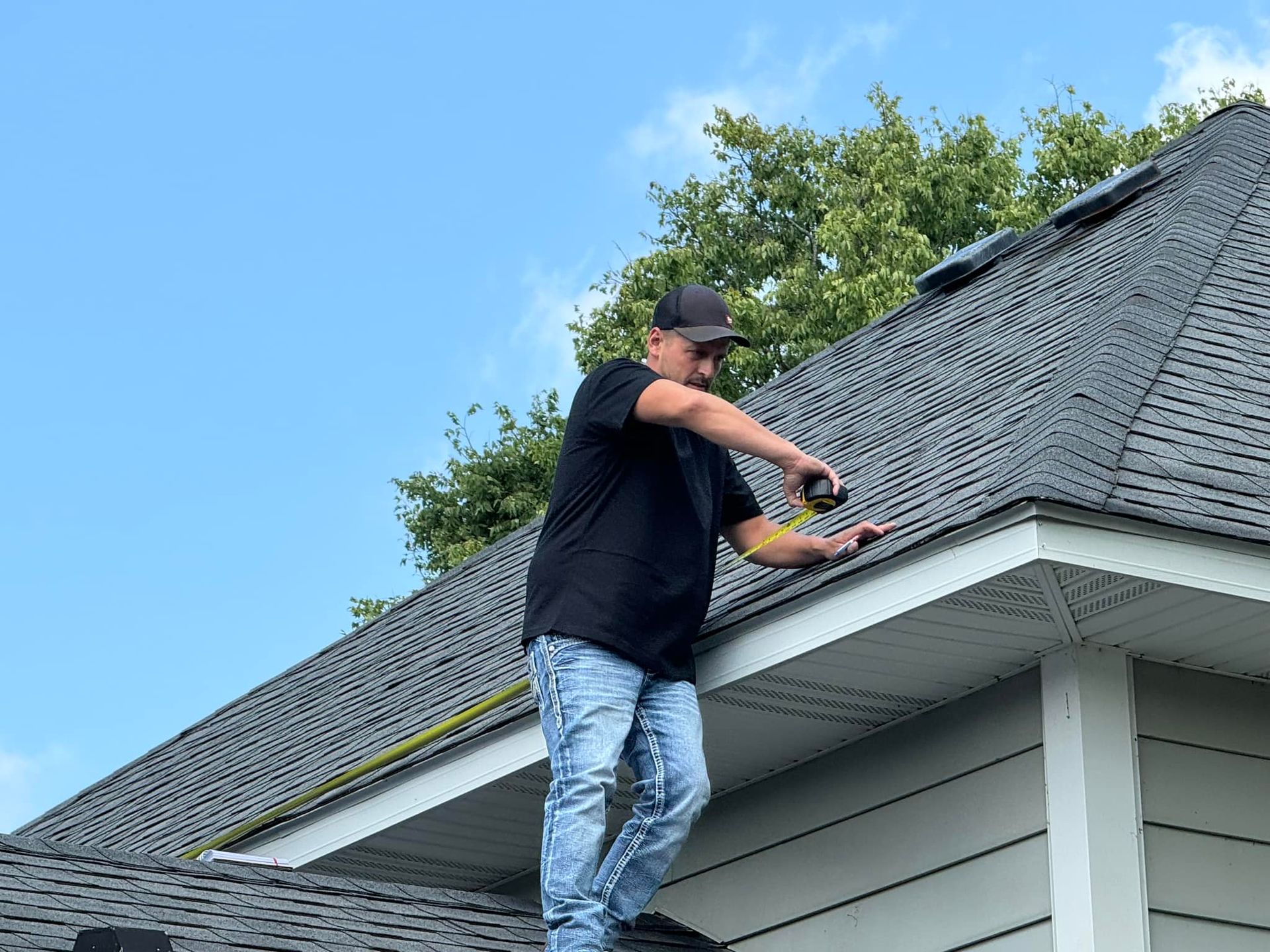 Person working on a gray shingled roof of a house, using a tool near the edge.