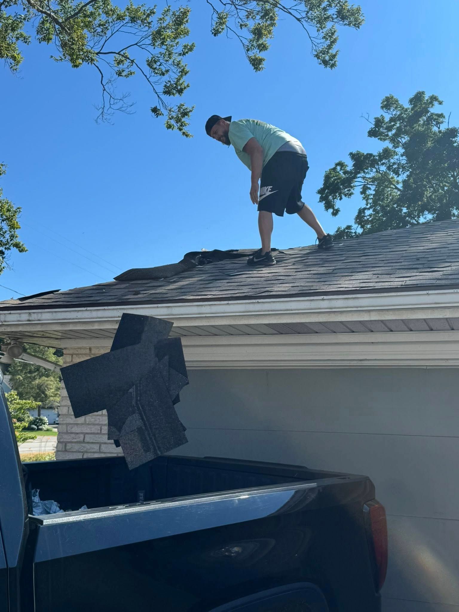 Person on a shingled roof near a tree, working above a parked truck under a clear blue sky