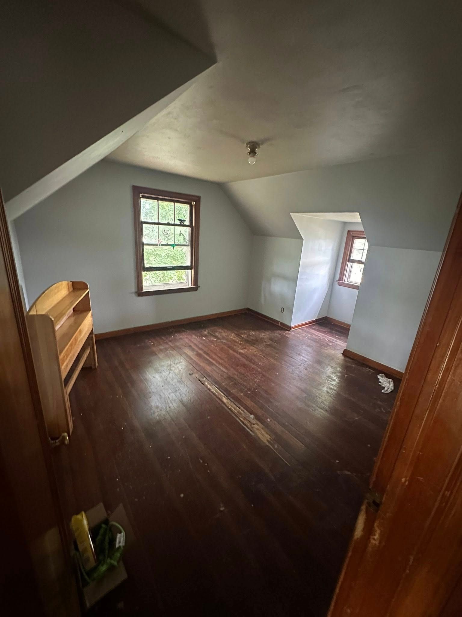 Empty attic bedroom with dark hardwood floors, sloped ceiling, and a window beside an open door.