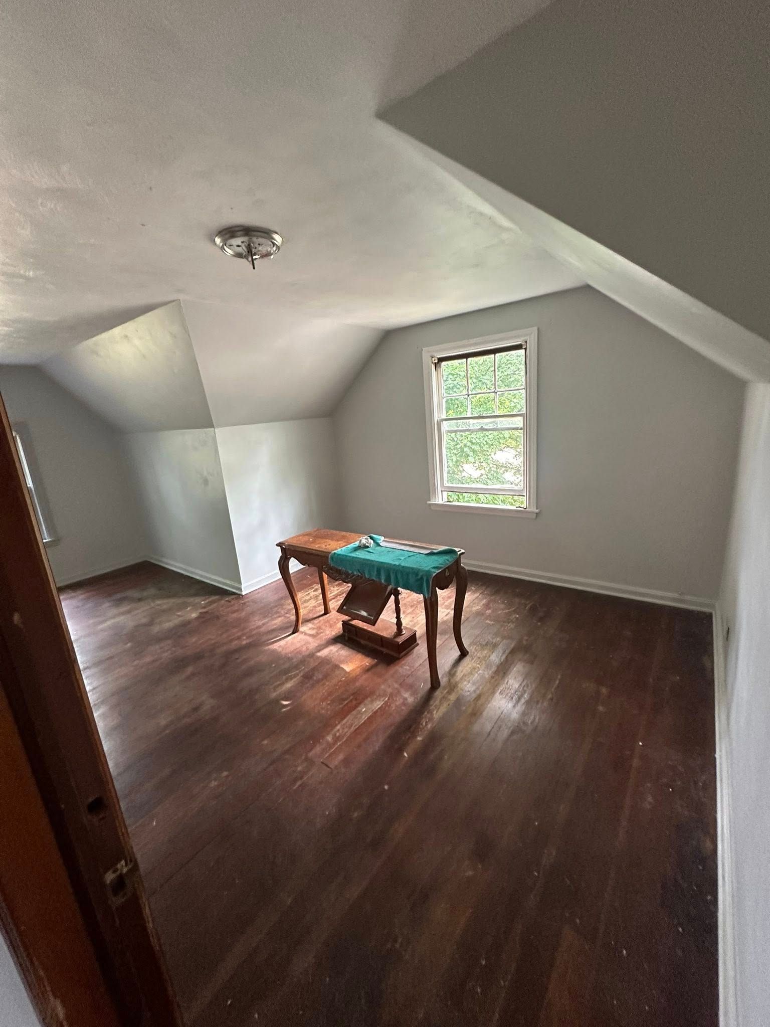 Empty attic room with dark wood floors, white sloped ceiling, and a small window with a table near it