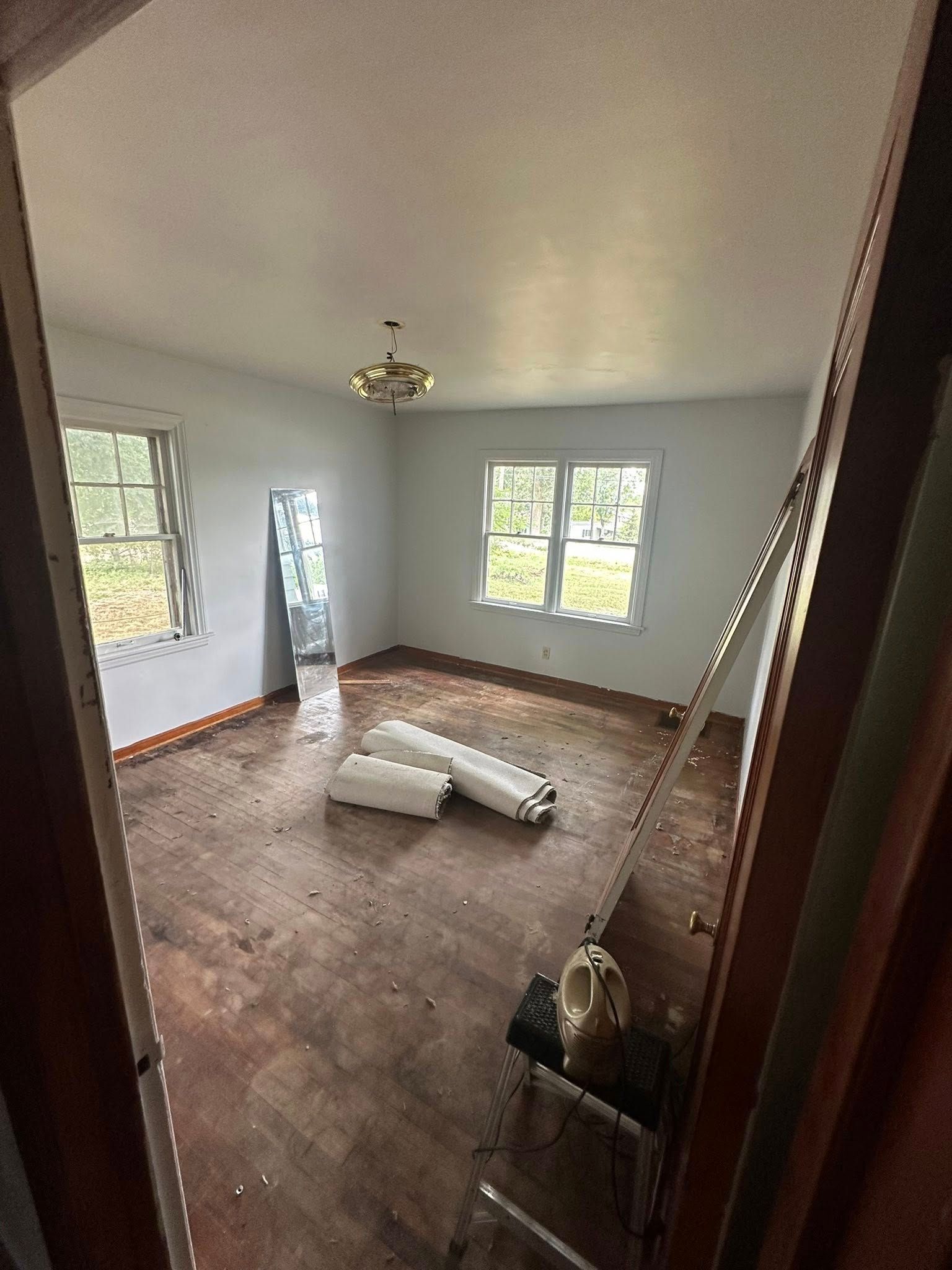 Empty room with white walls, hardwood floor, and scattered rolled materials near windows and a ceiling light.