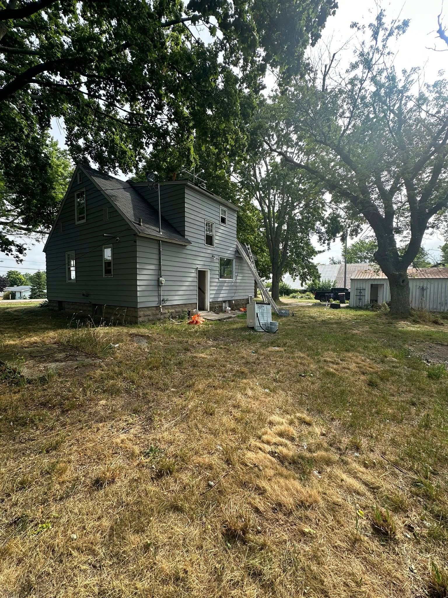 Small gray house under trees with a leaf-covered yard and a white shed in the background.