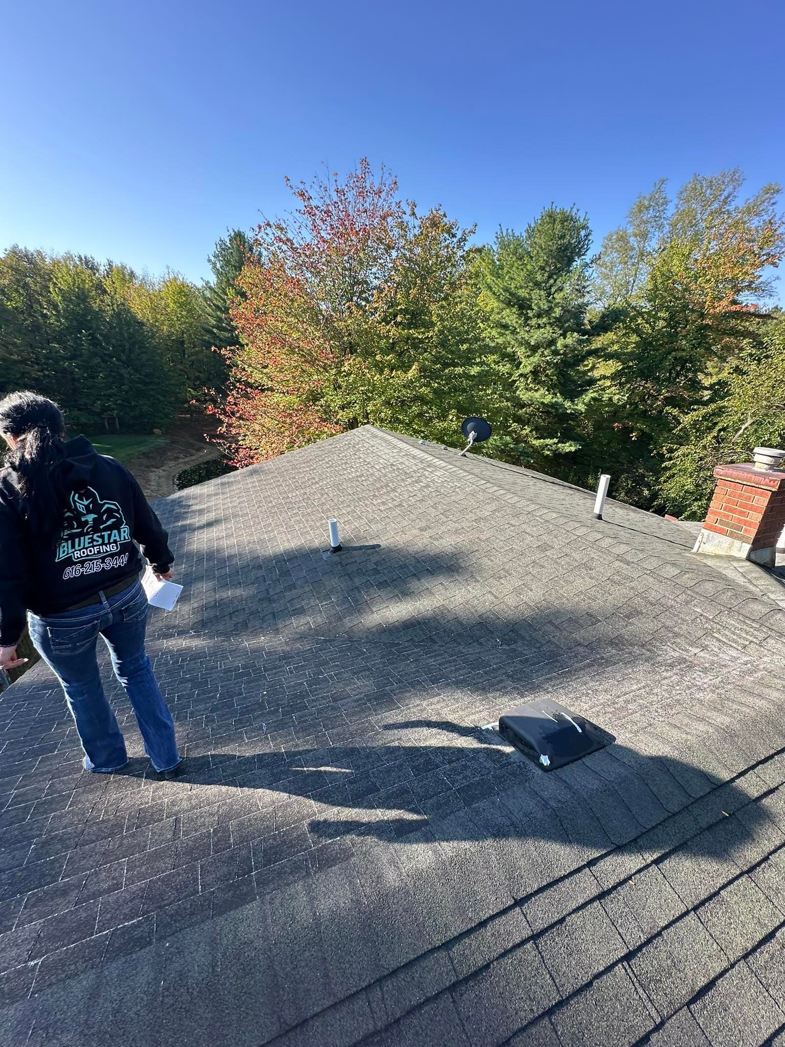 Person inspecting a gray shingled roof in bright sunlight with trees and blue sky in the background