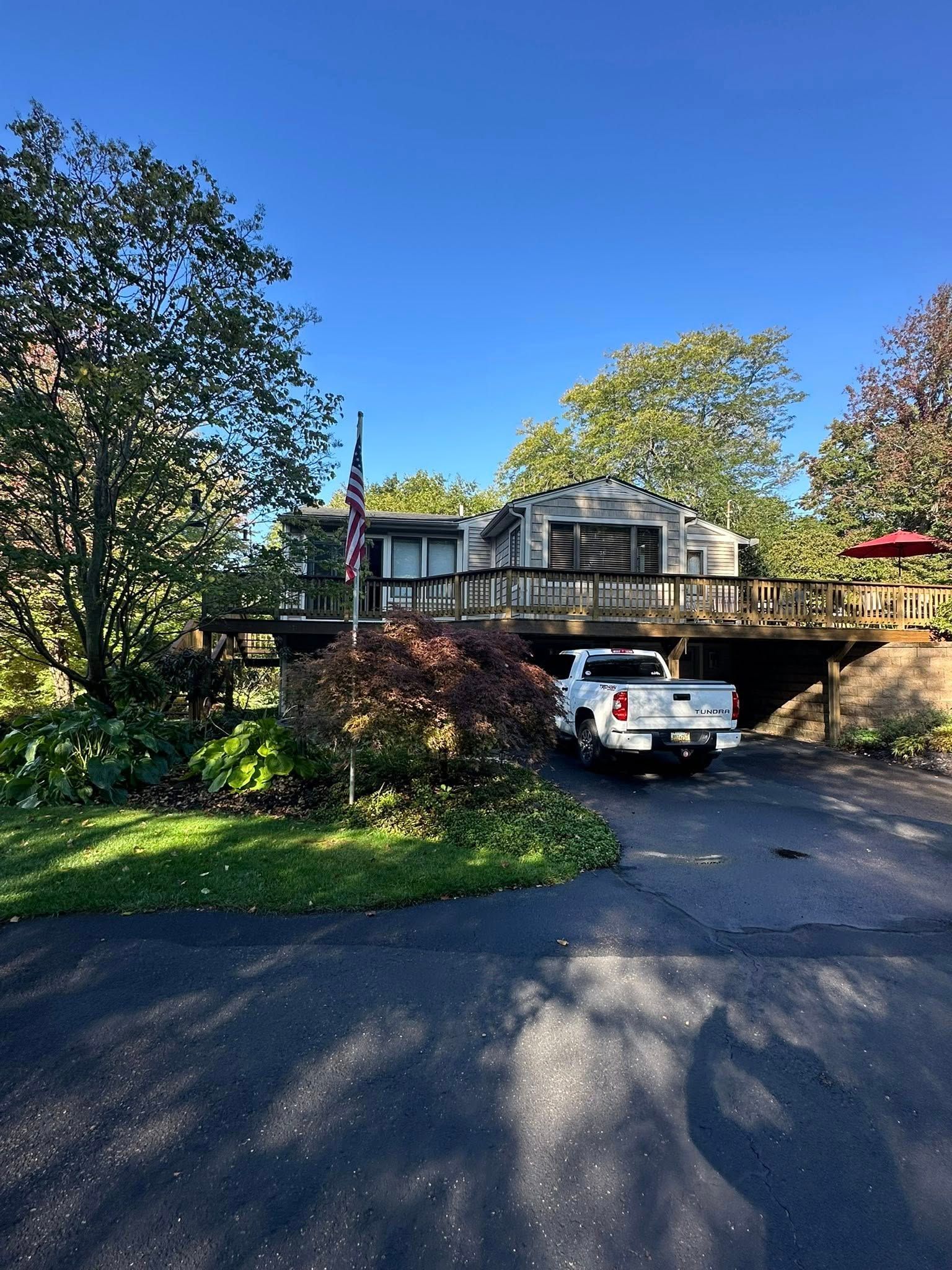 White car under a tree-lined driveway beside a wooden house with a porch in bright sunlight.