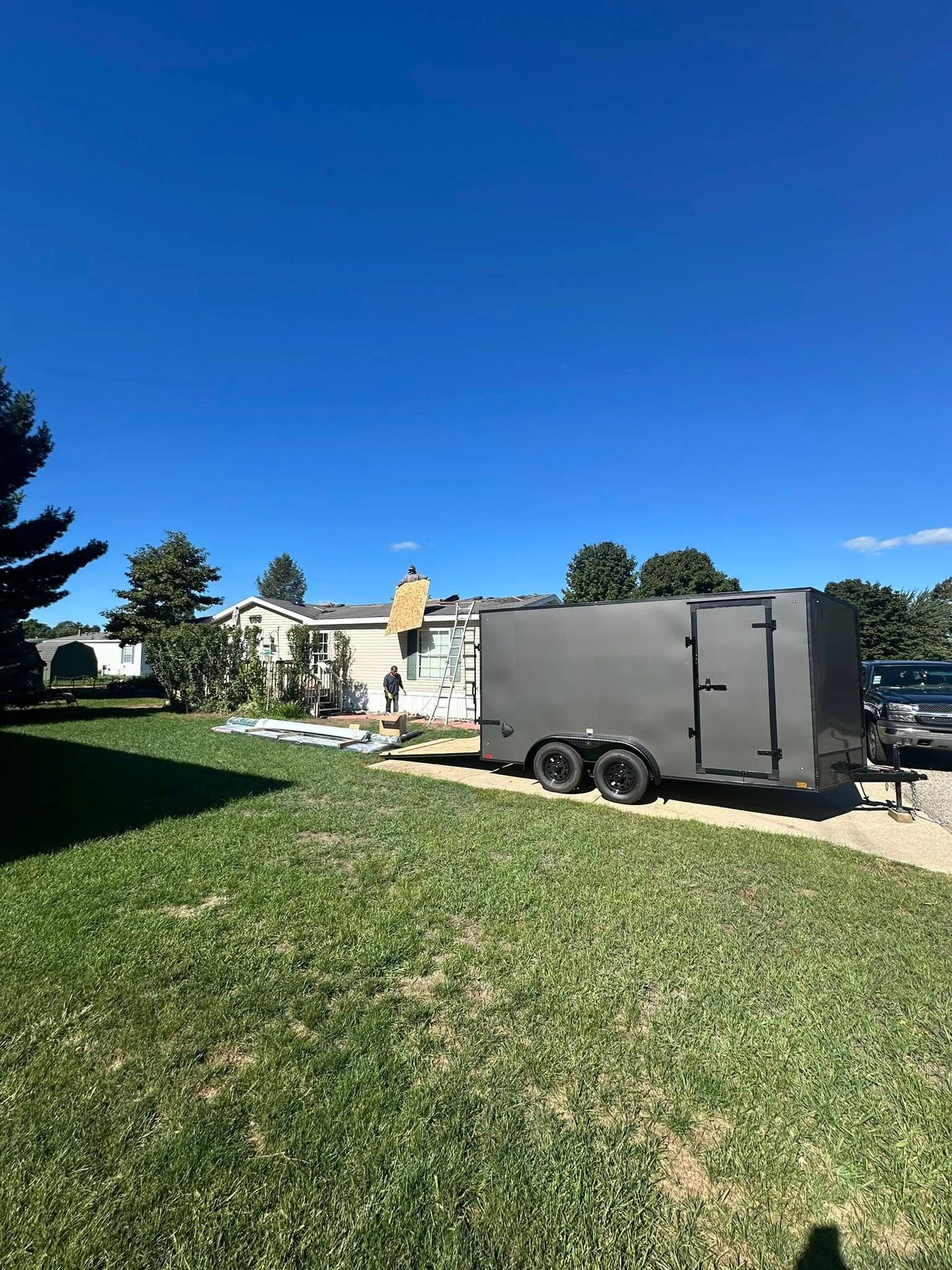 Gray enclosed trailer parked on a grassy driveway beside houses under a clear blue sky