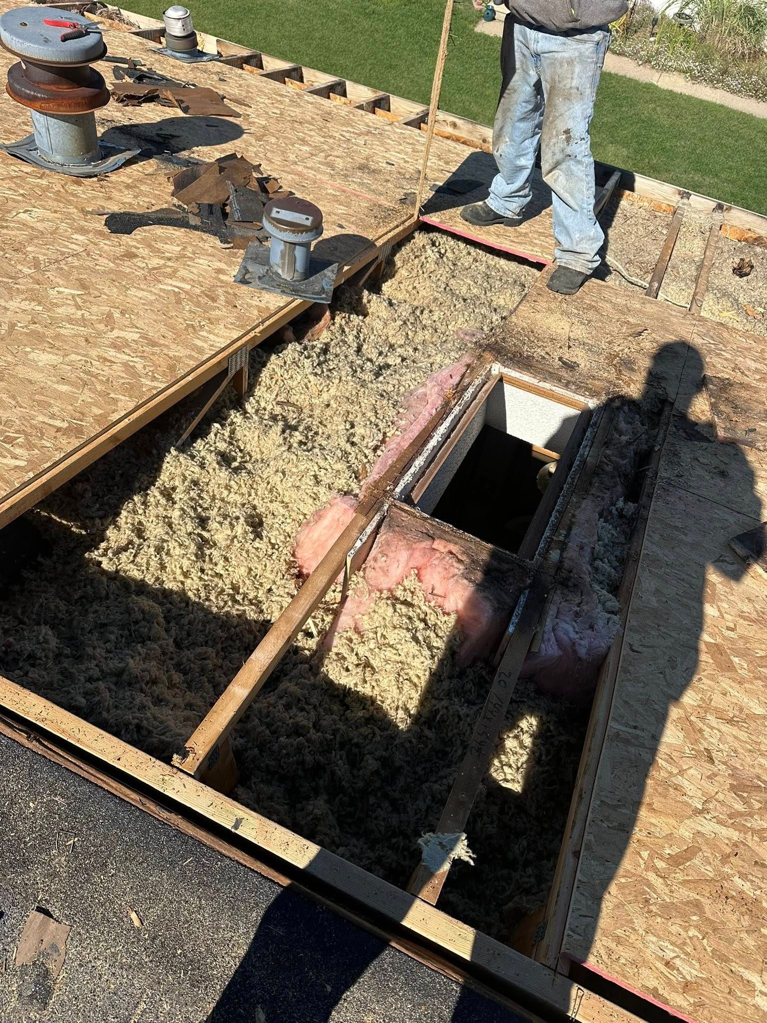 Roof under construction with exposed framing, gravel, and workers’ legs visible near an open hatch.