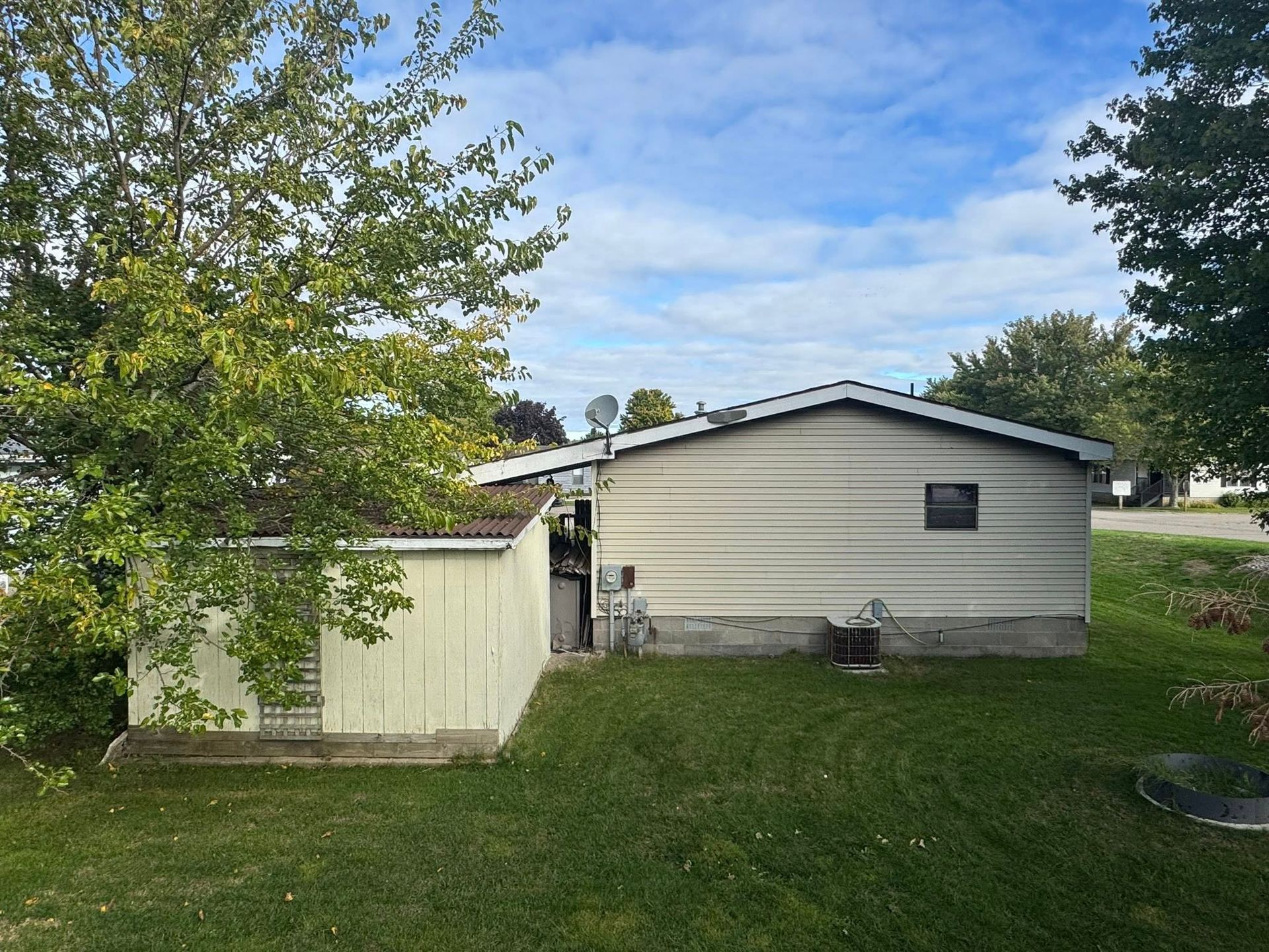Small beige house with a white shed in a grassy yard under a partly cloudy sky.