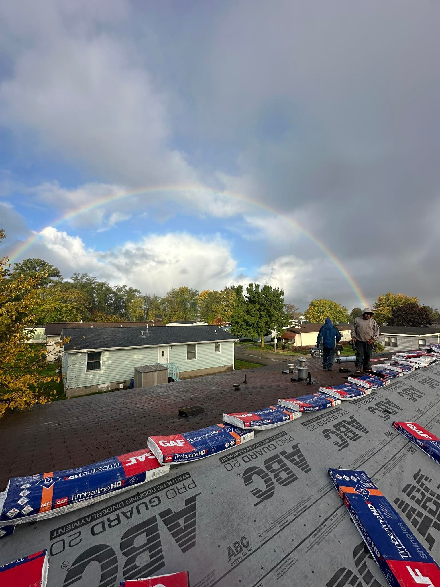 Roofing materials on a house under a cloudy sky, with workers on the roof and a faint rainbow in the background.