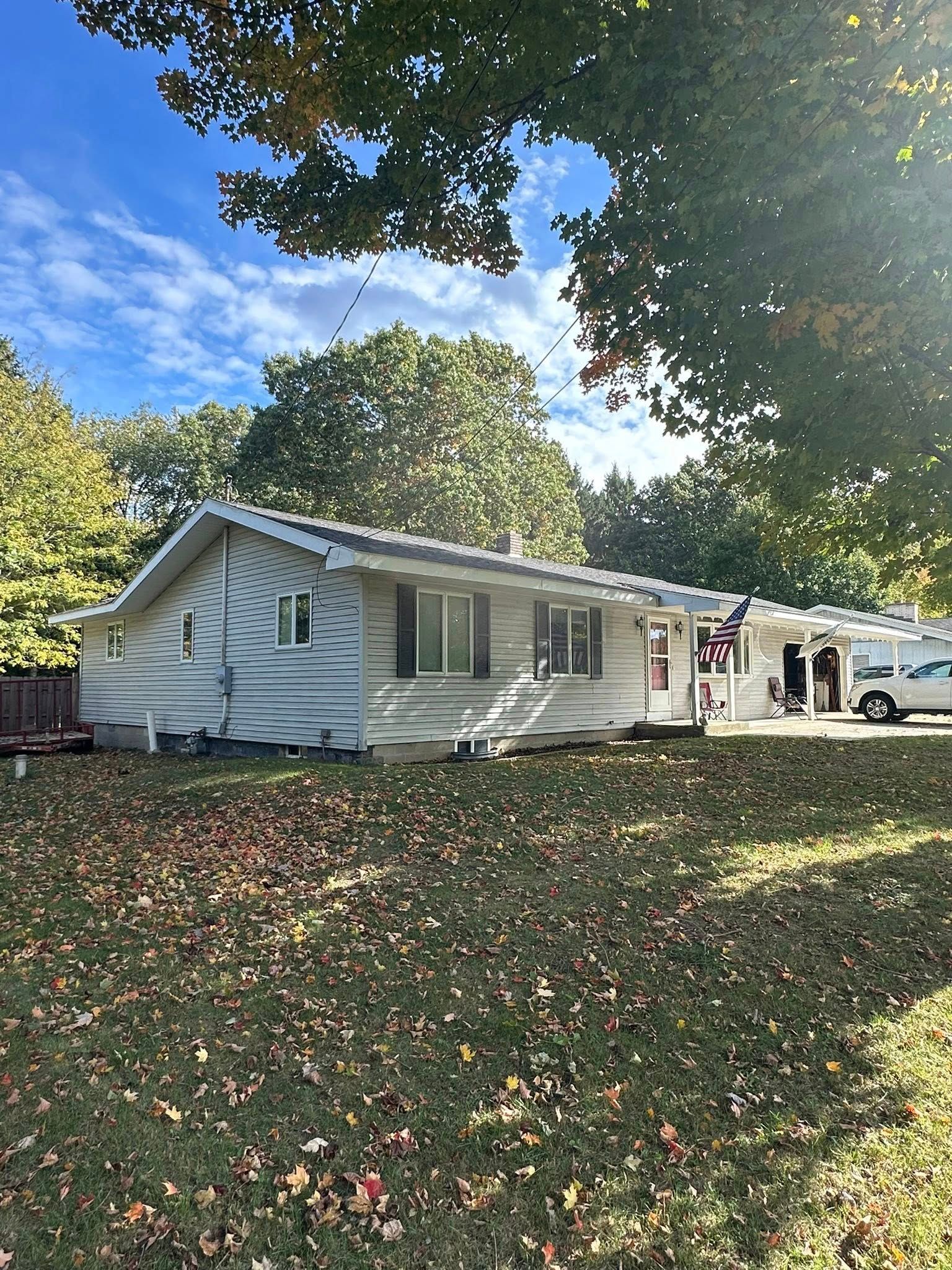 White house with a carport under large trees, surrounded by fallen leaves on a sunny day.