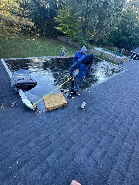 Worker in blue rain gear repairing a shingled roof near a skylight and chimney.