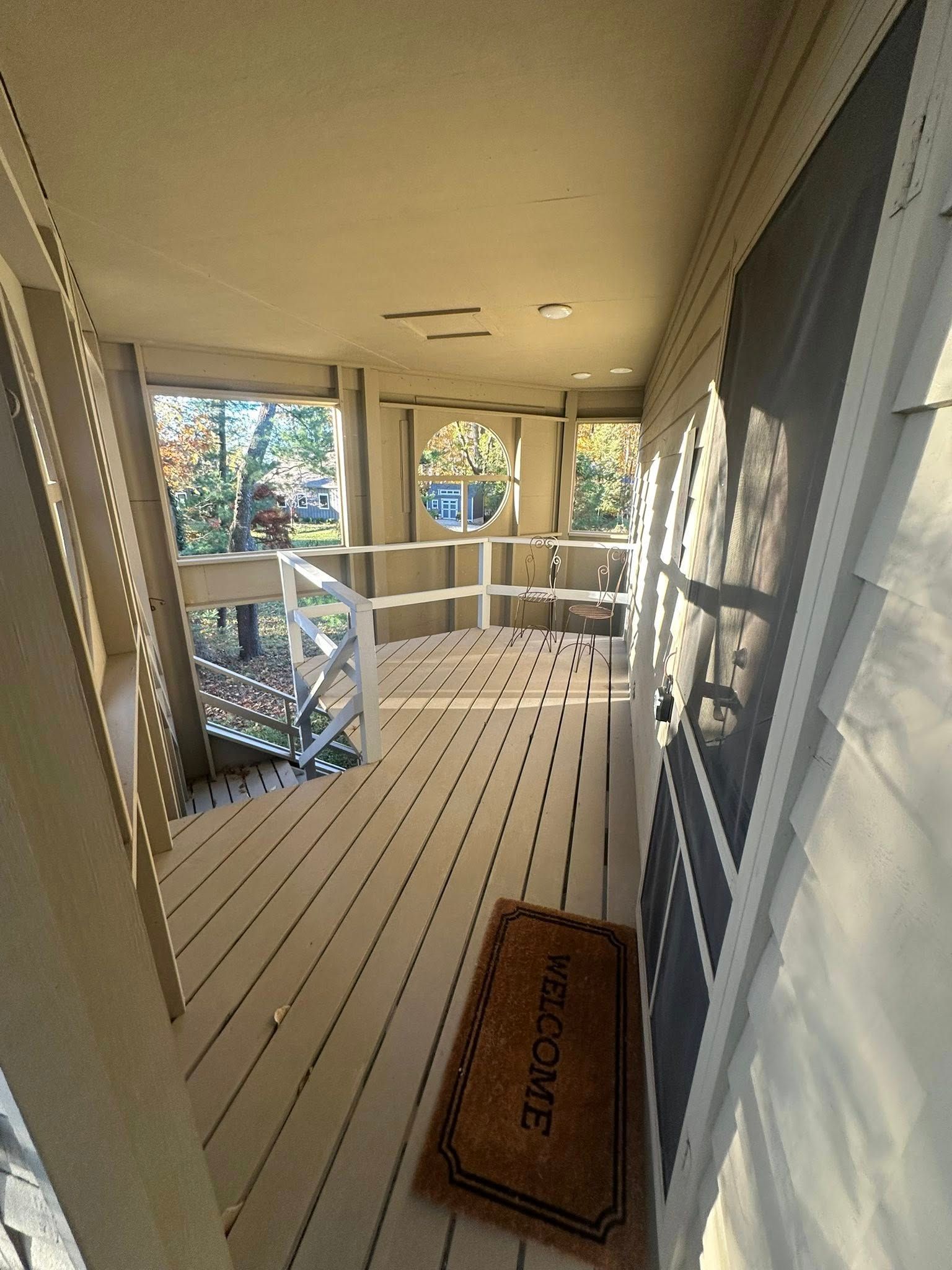 Covered porch with white floorboards, gray siding, and a welcome mat overlooking trees and a white railing.