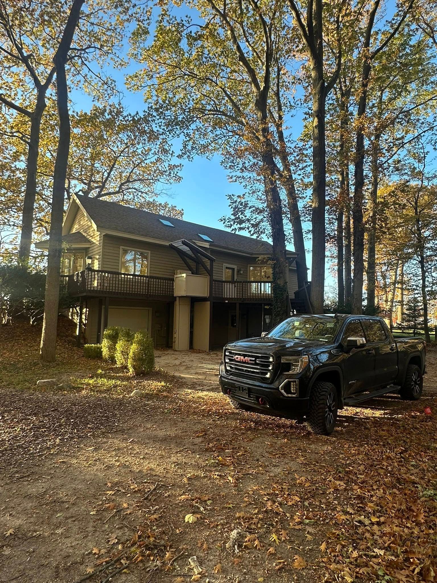 Black SUV parked beside a house in a wooded yard covered with fallen leaves.