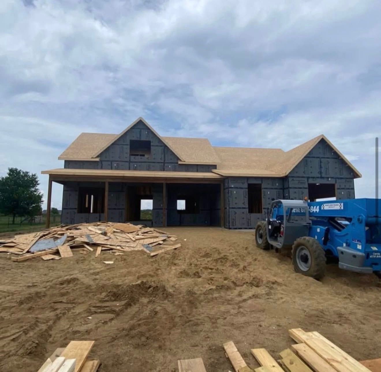 New house under construction with blue lift, wood framing, and lumber piles on muddy lot