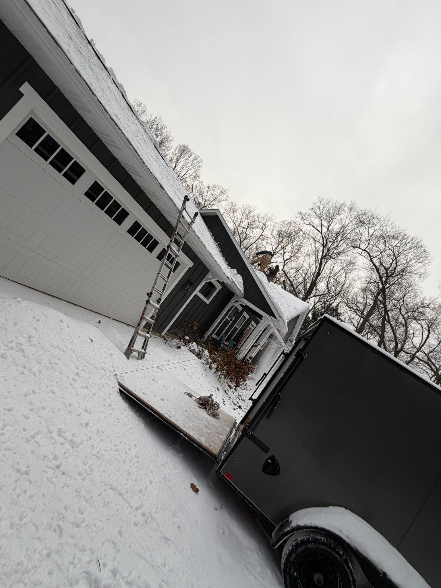 Snowy driveway beside a garage and black trailer under an overcast sky