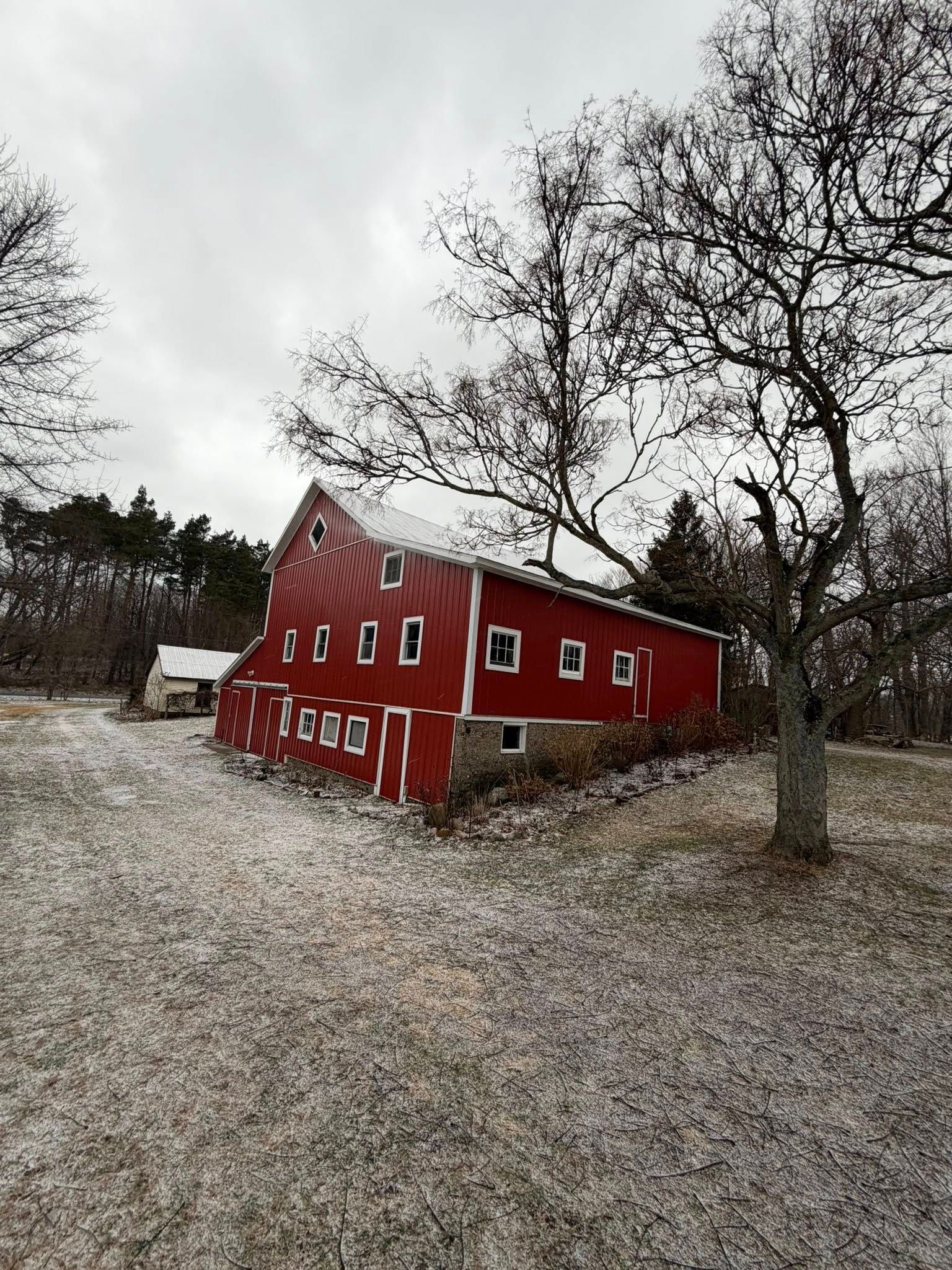 Red barn leaning in a snowy field beside bare trees under an overcast sky